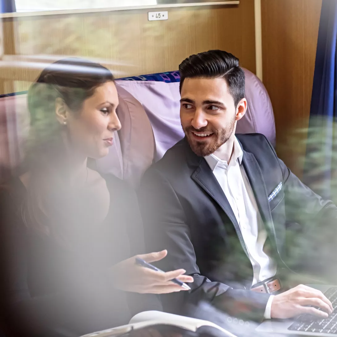 A woman and a man with a laptop are sitting in a train compartment and discussing